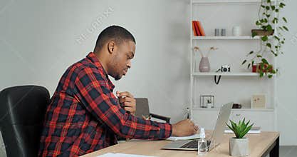 Busy african american guy student doing homework for university at home using laptop and notebook, focused business man