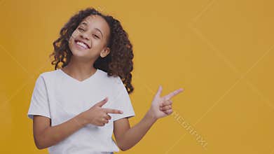 Cute african american teen girl pointing aside and smiling, orange studio background with empty space for mockup