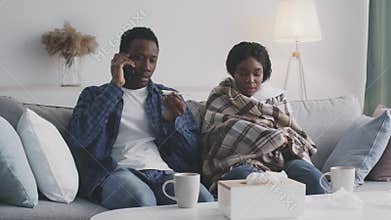 Black caring husband calling to doctor with cellphone, saying temperature of his sick wife, sitting on coach at home