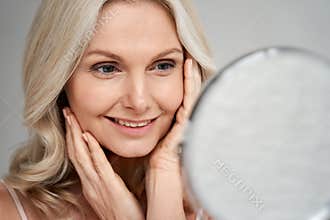 Happy 50s middle aged woman touching face skin looking in mirror.