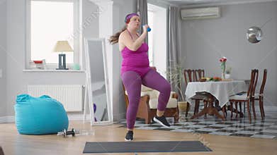 Wide shot portrait of positive obese Caucasian young woman singing dancing enjoying music in headphones exercising at