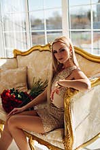 Stylish beautiful young blond girl fashion model wearing festive short dress posing sitting on couch with roses bouquet