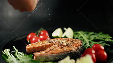 A cooked salmon steak with herbs and vegetables on a black serving plate is sprinkling spices. Grilled fish. Concept of