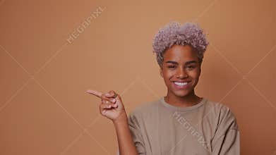 Dark skinned woman with appealing smile pointing on left side over brown wall.