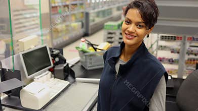 Young woman cashier posing for camera and smiling while sitting at checkout of store spbi.