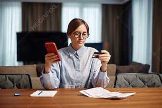 Focused young business woman in glasses pay bills or debt loan online on phone with documents sitting at desk. Ecommerce