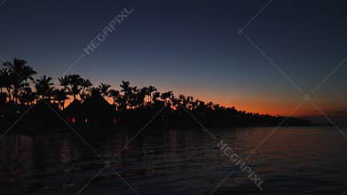 Tropical island at sunset, palm trees and beach bungalow. Romantic night, glowing lights over the sea water.
