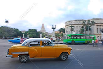 Classic yellow vintage Cuban car fast speeding with blurred background of yank tank tanks, bus & Capitol National at Havana, Cuba