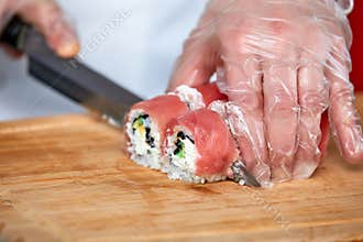 Chef cuts sushi rolls with red salmon fish with a knife on a wooden board. Chef in transparent gloves works.