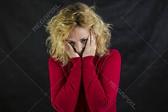 Middle-aged blond woman with red sweater on a dark background holds her head in her hands in despair
