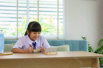 Asian elementary school student girl in uniform eating breakfast cereals with milk and looking cartoon in smartphone to