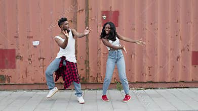 Young dancers on the street. Black guy and girl make the same movements while dancing. They stand next to high fence.