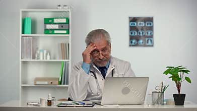 Smart male doctor in white medical coat using laptop for remote patient consultation in hospital office. Doctor talking
