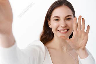 Young woman saying hello via video call, holding smartphone and taking selfie as waving hand and smiling friendly