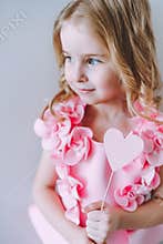Portrait of little blonde smiling girl holding pink wooden heart in front of her face.