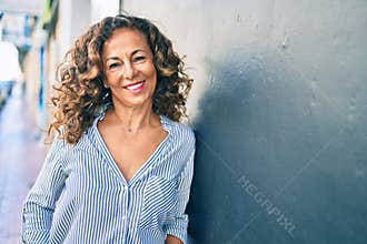 Middle age hispanic woman smiling happy leaning on the wall at the city