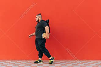 Full-length portrait of a stylish fat man in casual clothes walking with a backpack on his back and notebooks in his hands on a