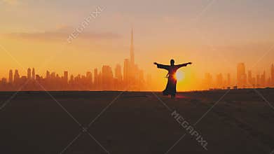 Arabic woman weared in traditional UAE dress - abayain rising her hands on the sunset at a desert with Dubai city