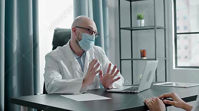 Bald doctor in white coat and mask types on laptop in office
