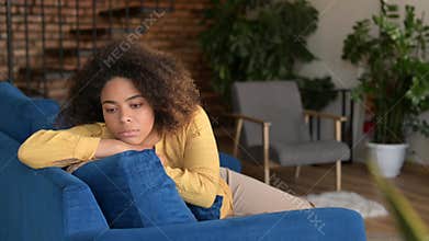 Upset African-American woman feels bored sitting on the couch at home alone