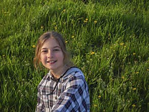 Smiling teenage girl on a walk in the park sits on a blooming green meadow in the sun