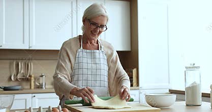 Happy blonde senior baker woman in kitchen apron rolling dough