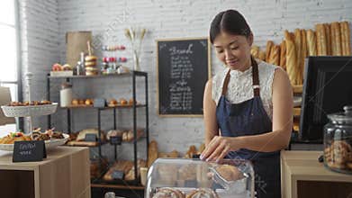 Beautiful woman working in a bakery shop arranging bread and pastries while smiling at the counter
