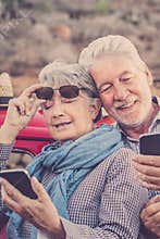 Cheerful old senior couple use phones together outdoor with red car in background - concept of travel mature people and modern
