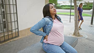 Tired young pregnant woman aching from backache, sitting on city street bench, touching her belly in thoughtful contemplation of