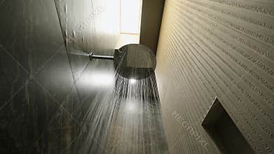 Bottom view of water running from round silver shower head in stylish interior with dark tiles on wall. Luxury bathroom
