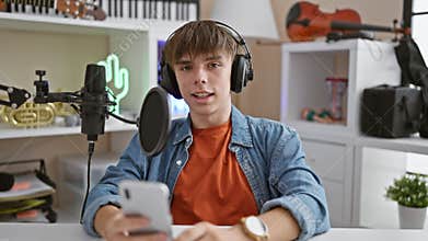 Young caucasian man podcasting from home with microphone, headphones, smartphone, and musical instruments in the background