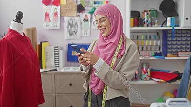 Smiling woman in hijab using smartphone in a colorful tailor shop with mannequin and fabrics