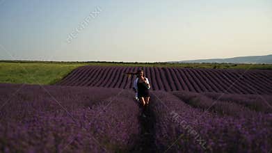 A woman is runs through a field of purple flowers. She is wearing a white shirt and a hat.