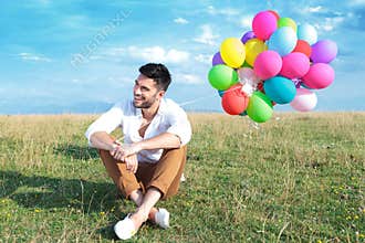 Casual man with balloons sits on grass and smiles