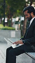 Bearded businessman opening pc and typing while sitting outdoors