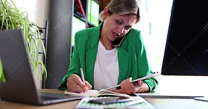 Serious business woman talking on mobile phone at workplace in office making notes and working at computer
