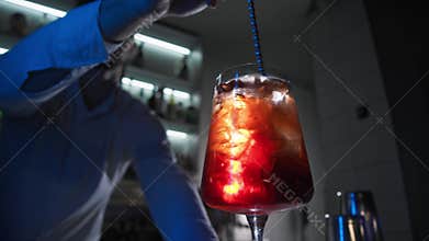 Barman in nightclub stands behind bar and prepares drink. He mixes ice with spoon in glass, creating delightful Aperol