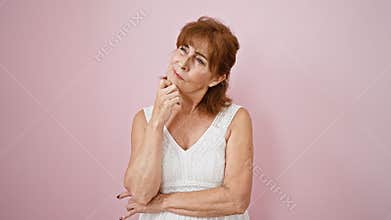 Pensive middle age woman in dress on pink wall background, hand on chin, lost in thought, guessing over a question. doubting with