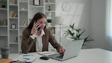Focused Businesswoman Speaking on the Smartphone While Sitting at Home Office and Using the Laptop.