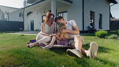 American dream family spends a free warm summer day outdoors near their house, sitting on the lawn. Mom, dad and