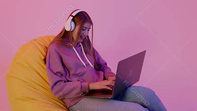 Youth girl in hoodie and jeans lying on bean bag with notebook. Youngster in wireless headphones talking with her