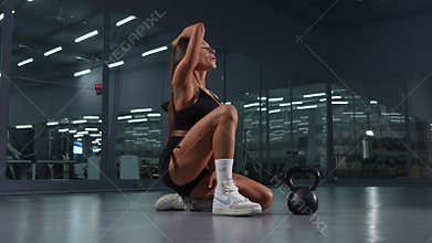 A beautiful brunette with large breasts sits on the floor in the gym. Caucasian woman posing with kettlebell.