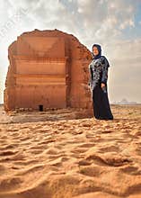 Woman in long dark dress posing in front Mada\'in Salih Lonely Castle or Qasr al-Farid at Hegra, Saudia Arabia, sandy desert