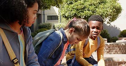 3 students seeing sad peer, leaning in, hugging to comfort on brick bench at school backpacks
