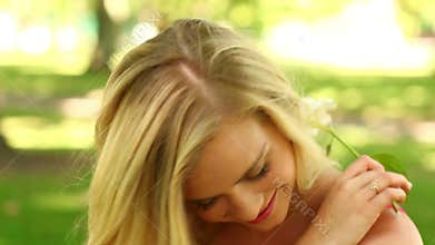 Pretty blonde relaxing in the park smiling at the camera while smelling a flower