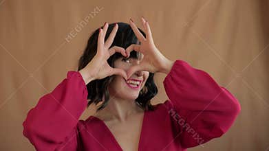 Playful woman forming heart shapes with hands near eyes and smiling in fashion red blouse. Gesture and wide happy smile convey