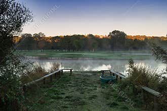 Morning fog settles over a calm lake surrounded by trees and greenery