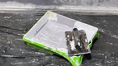A close-up reveals a paint tray with gray paint and a spatula, set on a plastic sheet, ready for home improvement.