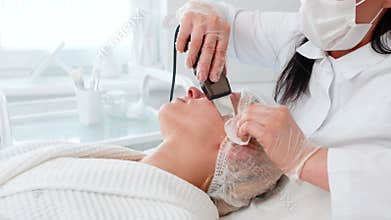Cosmetologist using ultrasonic scrubber device for hardware facial cleansing on middle aged woman