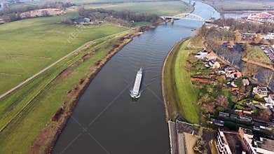Aerial view of river with cargo ship, arched bridge, and mixed use banks. Residential homes, gardens, and green fields frame a